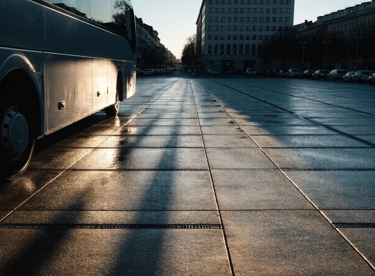 Eye-level view of a Prague bus terminal departure apron in cold morning light, coach flank at left, slab tarmac with long shadows