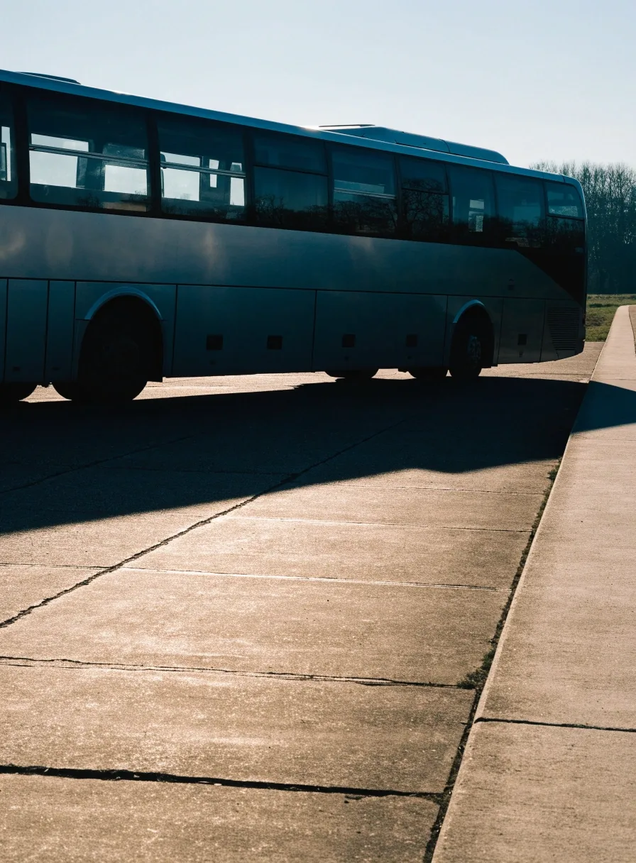 Regional coach parked at a terminal apron in cold morning light, slab tarmac and long shadows, unbranded coach flank